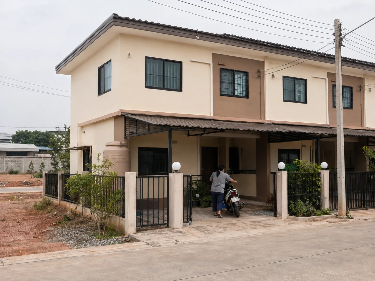 ภายนอกบ้านทาวน์เฮาส์ของรากฐาน ผนังสีครีม ที่จอดรถมีหลังคา แนวหน้าบ้านเล็ก ๆ ใต้ท้องฟ้าครึ้ม